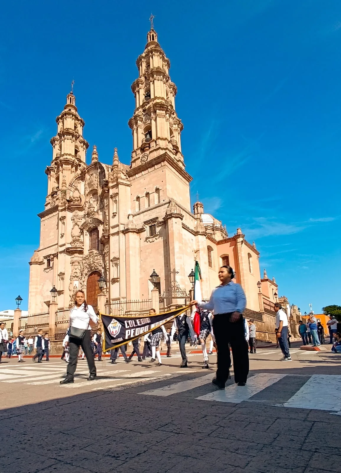 La comunidad de Primaria desfila detrás del banderín del Colegio Pedro Moreno frente a un templo de dos torres.